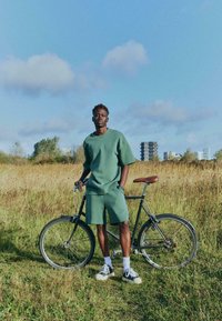 Homme en short et chemise verts debout avec un vélo noir dans un champ herbeux sous un ciel bleu avec des bâtiments de la ville en arrière-plan.