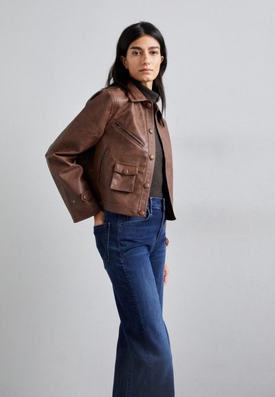 Chaqueta de cuero marrón con un diseño ajustado, que presenta un cierre de botones, dos bolsillos frontales y un bolsillo en el pecho con cremallera. Combinada con jeans azul oscuro.