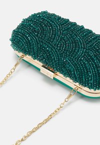 Tajemnicza torebka na koralikach z złotym okuciami, z wzorem fal. Zawiera odłączany złoty łańcuch na ramię. Zwarta prostokątna forma.