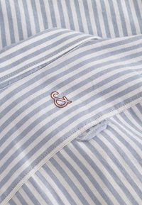 Tessuto a righe blu chiaro e bianco con una consistenza morbida, caratterizzato da un piccolo ricamo del logo color maroon. Cuciture lisce lungo i bordi.