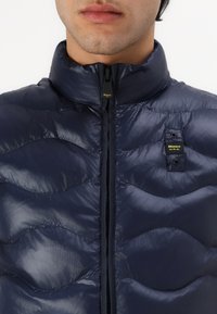 Marineblå dunjakke med høy krage, blank finish, horisontal quiltet mønster, full front glidelås og logo-patch på brystet.