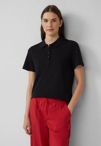 Polo shirt noir à manches courtes avec col et patte de boutonnage à cinq boutons, associé à un pantalon rouge ample. Tissu lisse, design décontracté.