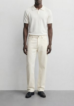 Muž v krémovej polo košeli s krátkym rukávom, svetlo béžových rovno strihaných nohaviciach a čiernych mokasínach, stojaci pred jednoduchým svetlým pozadím.