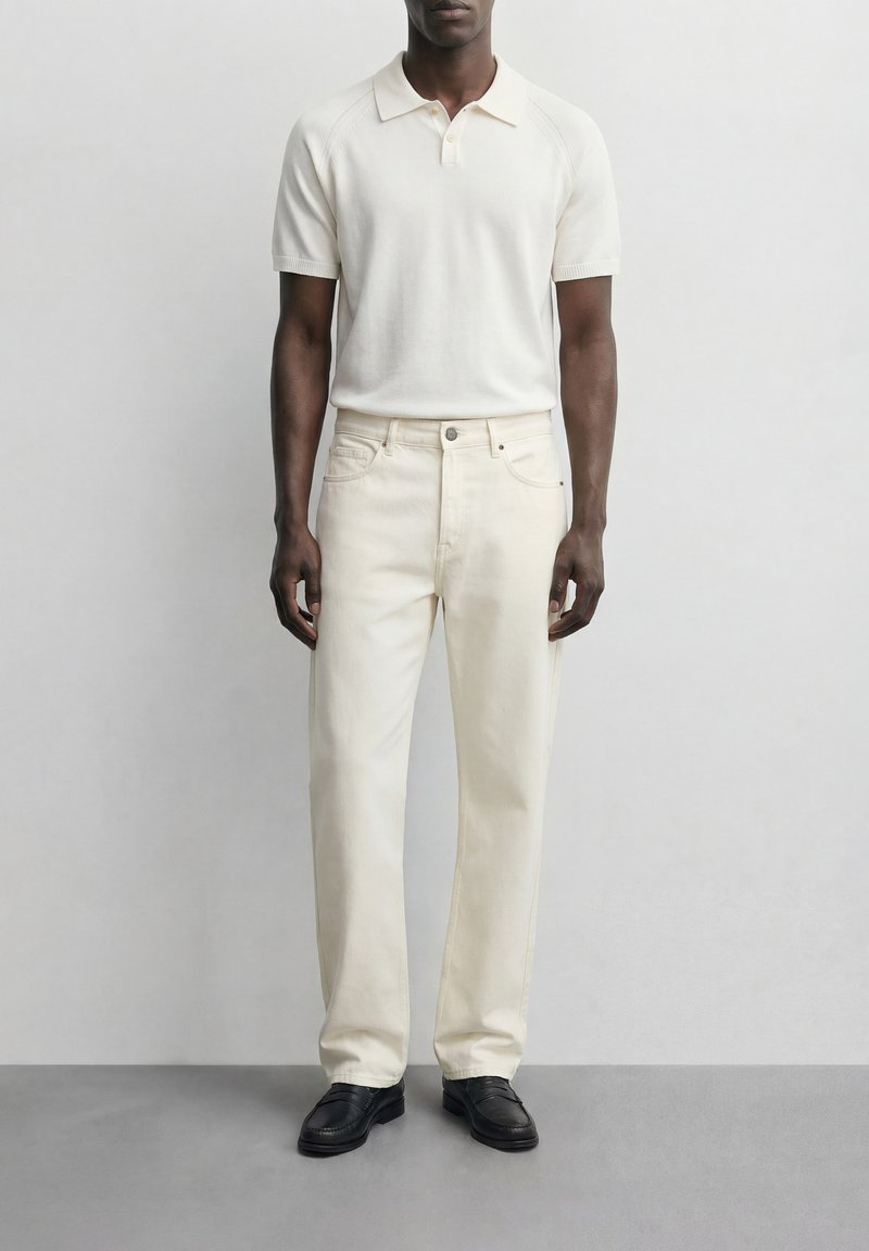 Άνδρας που φοράει κρεμ κοντομάνικη πόλο μπλούζα, ανοιχτό μπεζ παντελόνι ίσιας γραμμής και μαύρα μοκασίνια, στέκεται μπροστά σε ένα απλό φωτεινό φόντο.