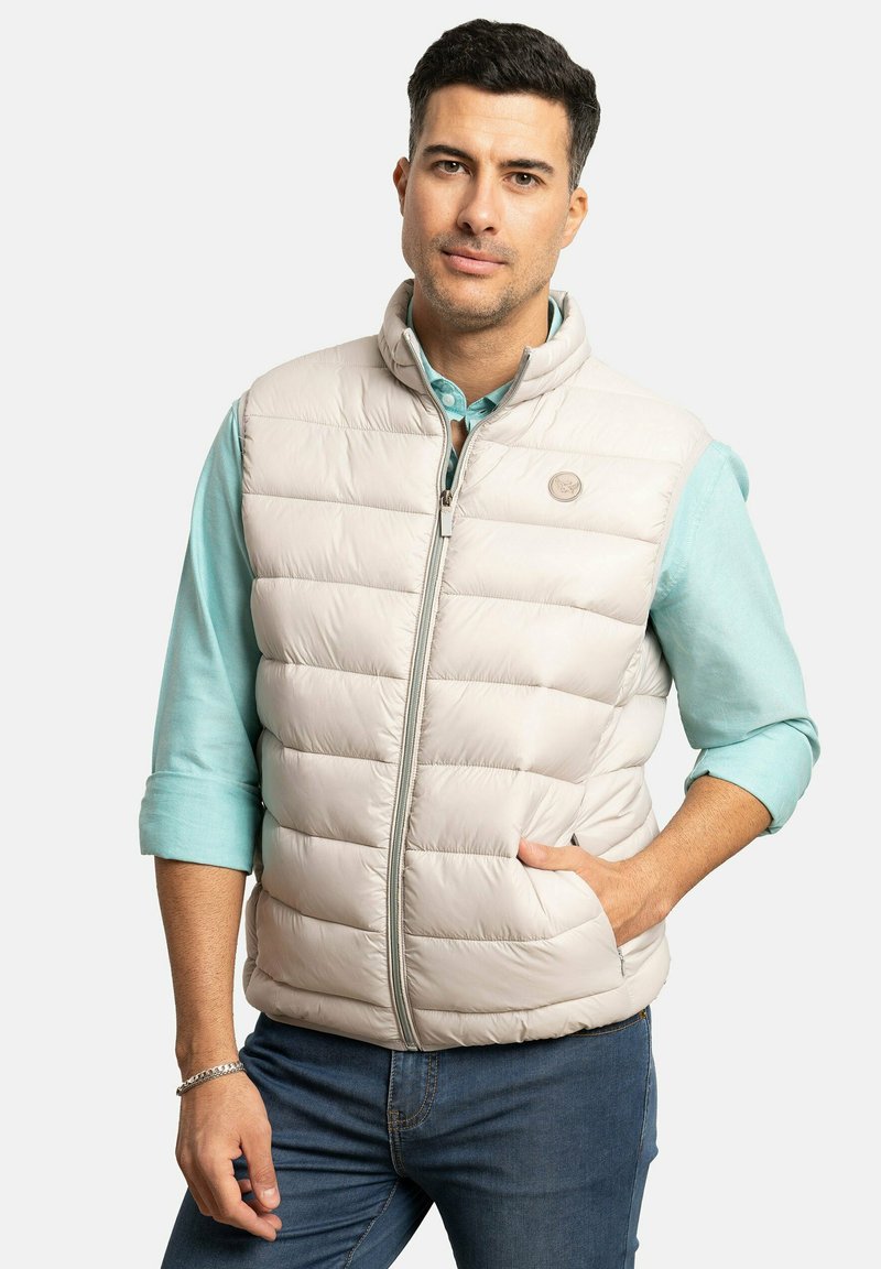 Chaleco acolchado de color beige claro con cremallera frontal y bolsillos laterales. La tela parece suave con un brillo sutil. Logo redondo en la parte superior izquierda del pecho.