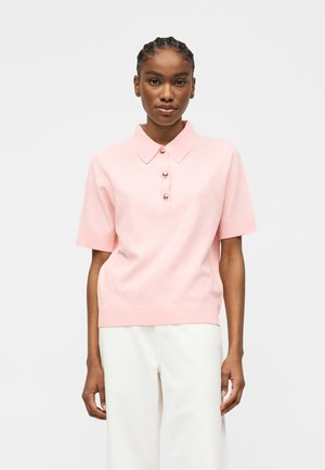 Femeie cu părul împletit, purtând un tricou polo roz deschis cu mâneci scurte și pantaloni albi, stând în picioare în fața unui fundal simplu.