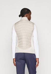 Lys beige polstret vest med et glatt design og kviltet tekstur, parret med en tettsittende hvit langermet topp og mørke bukser.