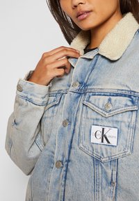 Vaaleansininen farkkutakki, jossa on tekoturkistakki, hopeiset yksityiskohdat, etutaskut ja näyttävä "Calvin Klein Jeans" -logo.