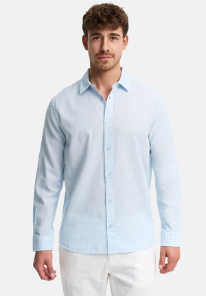 Młody mężczyzna z kręconymi włosami, ubrany w jasnoniebieską koszulę zapinaną na guziki i białe spodnie, stojący na tle jednolitej białej ściany.