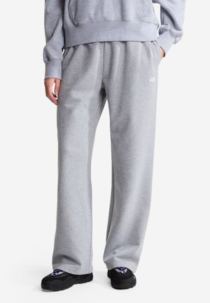 Person trägt hellgraue Jogginghose und passendes Sweatshirt, steht mit einer Hand in der Tasche, trägt schwarze und violette Sneakers.