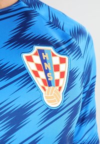 Jersey deportivo azul con patrones oscuros en zigzag y una insignia de fútbol croata que presenta cuadrados rojos y blancos, las letras HNS y un balón dorado.