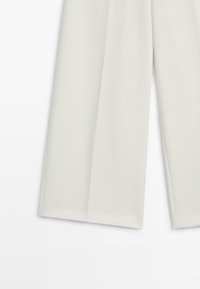 Pantalon à jambes larges de couleur blanc cassé, confectionné dans un tissu lisse. Il présente des plis marqués sur le devant et une silhouette fluide.