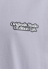 Světle levandulové tričko s černou grafikou na přední straně, která zní "Originals Studio © Edition Cph". Hladká bavlněná textura.