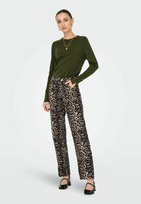 Grünes Longsleeve-Oberteil mit Knöpfen an den Manschetten, kombiniert mit beigen Hosen im Leopardenmuster. Schwarze High Heels ergänzen das Outfit.