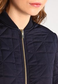 Chaqueta bomber acolchada navy con un patrón de diamantes, cremallera dorada y cuello acanalado. La textura del tejido suave realza su diseño moderno.