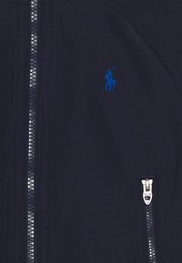Marineblauwe jas met zilveren ritsen en een klein geborduurd logo van een blauwe polo-speler op de borst.