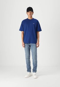 Blue cotton t-shirt with short sleeves and a round neckline, paired with light blue jeans and white sneakers. Black cap on head.