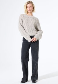 Vrouw met kort blond haar, gekleed in een beige kabelgebreide trui, zwarte wijde jeans en zwarte schoenen, staand tegen een witte achtergrond.