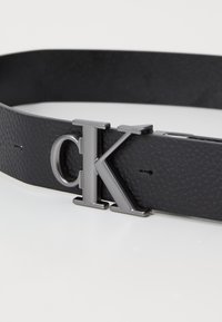 Črn usnjeni pas z teksturirano površino, ki ima kovinski sivo "CK" sponko za zapiranje, zasnovan v minimalističnem slogu.