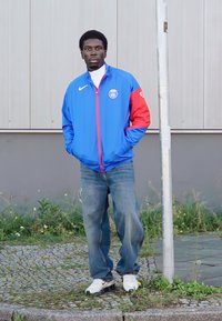 Kék Nike dzseki piros kiegészítőkkel, logóval, laza kék farmerral és fehér sportcipővel, semleges háttér előtt állva.