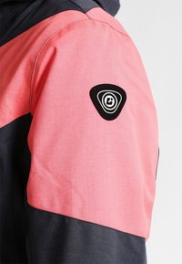 Personne portant une veste rose et gris foncé avec un patch logo triangulaire noir et blanc sur le haut de la manche.