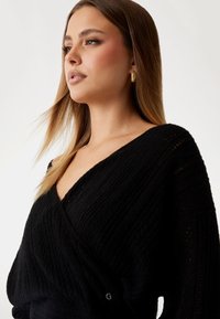 Noor naine, kellel on pikad heledat pruunid juuksed, kannab kuldseid rõngasKõrvaehteid ja musta tekstuuriga V-kaelusega kampsunit, vaatab vasakule.