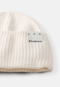 Valge soonikkoes beanie, volditud äärega. Omab ristkülikukujulist sildikest, millel on musta värvi tekst "CPH" ja "STUDIOS". Pehme tekstuur.