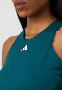 Teal Sporttop mit hohem Kragen, mit einem weißen Logo auf der Brust. Glatter Stoff, figurbetontes Design, ohne sichtbare Muster.