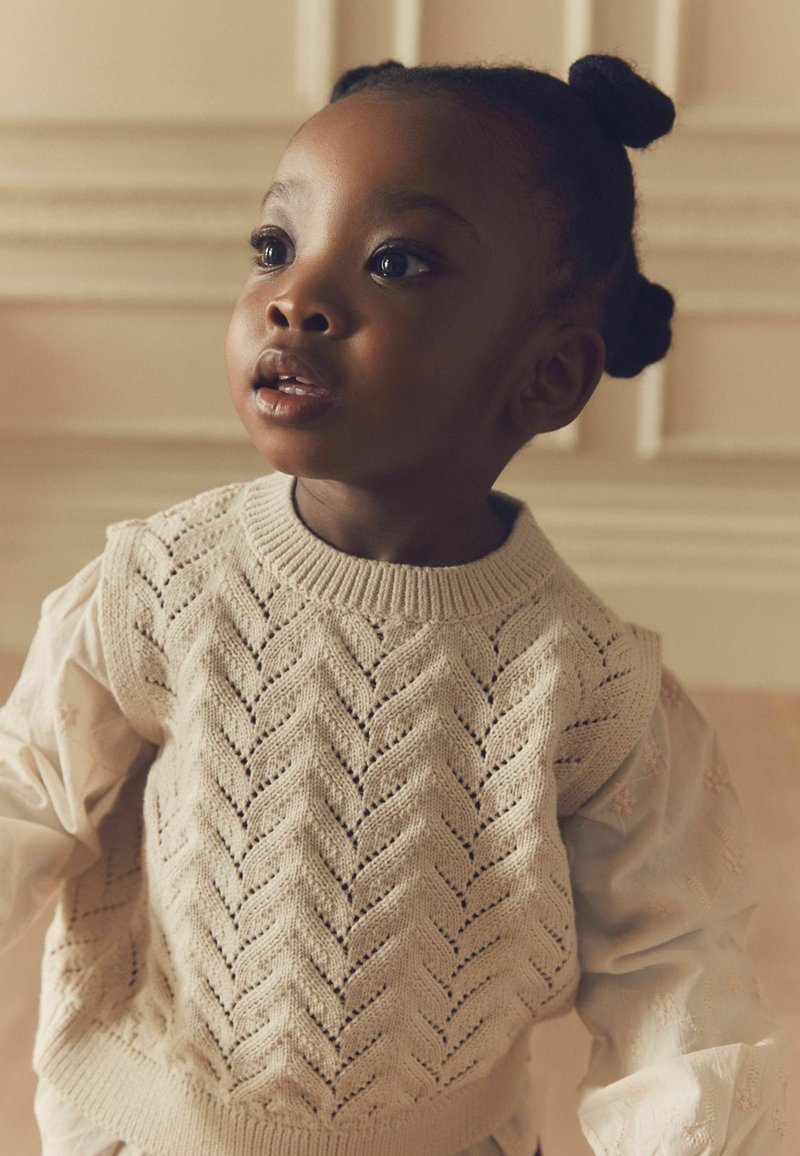 Striktrøje med et chevronmønster i lys beige, bæres over en lysefarvet, struktureret bluse. Barn med naturlig frisure og neutral baggrund.
