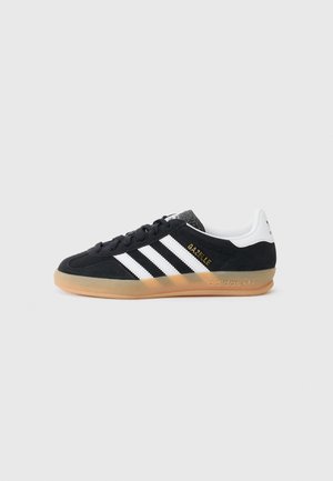 Baskets Adidas Gazelle noires avec des bandes blanches, semelle en gomme et languette de talon blanche, vues de profil sur fond gris clair.