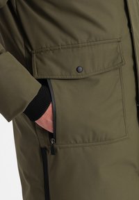 Chaqueta acolchada de color verde olivo con un gran bolsillo de solapa, un bolsillo con cremallera y puños de canalé en negro. Presenta una textura suave y un material ligero.