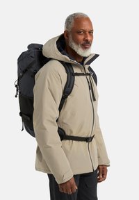 Giacca isolante beige con un grande cappuccio, zaino nero con pannello in rete, spallacci regolabili e chiusura zip sicura.
