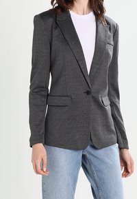 Blazer grigio in tessuto jersey con revers, chiusura a un bottone, due tasche frontali e vestibilità sartoriale; indossato sopra una maglietta bianca.
