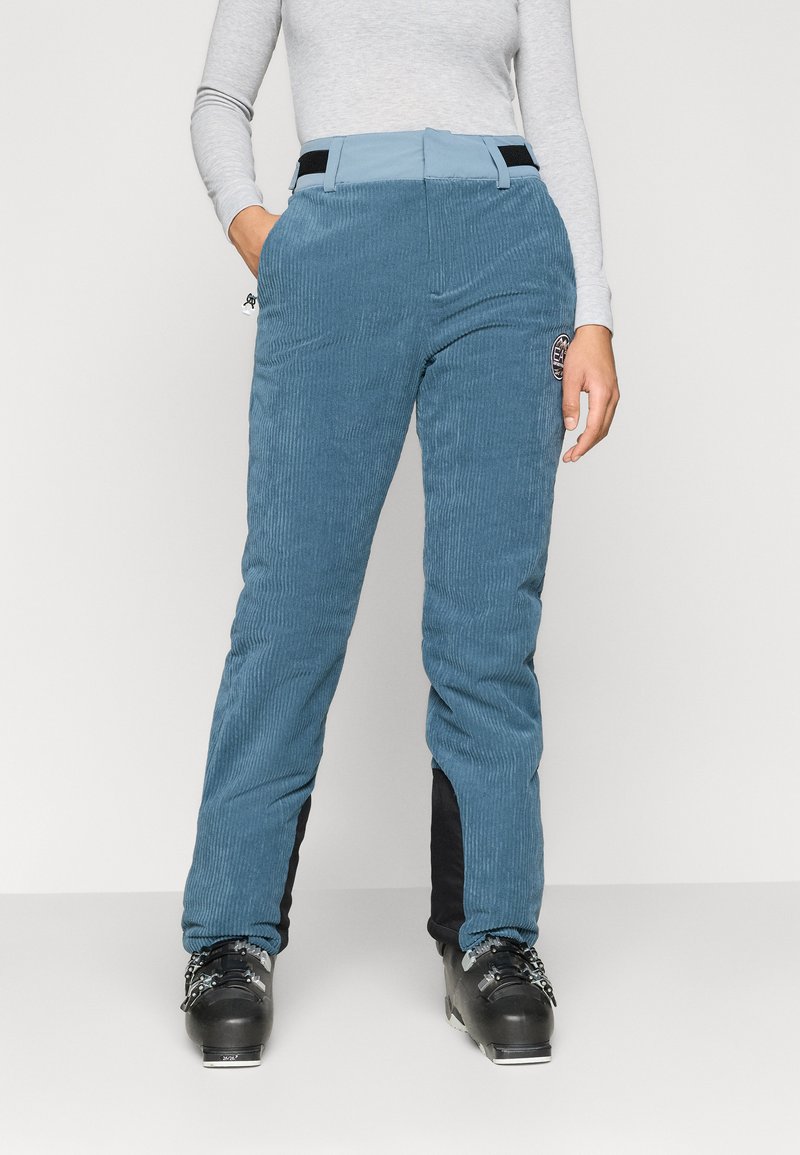 Calças de esqui em corduroy azul com painéis laterais pretos, apresentando uma cintura ajustada, bolsos laterais e um patch de logótipo na coxa. Botas de esqui pretas.