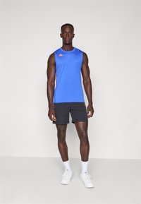 Blå ærmeløs sportstrøje med orange logo, parret med sorte shorts og hvide sneakers; glat stof og tætsiddende design.
