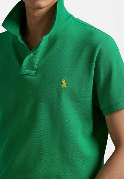 Zelena polo majica narejena iz teksturirane tkanine, z ovratnikom in z rumenim vezenim logotipom na levem prsnem delu. Kratki rokavi.