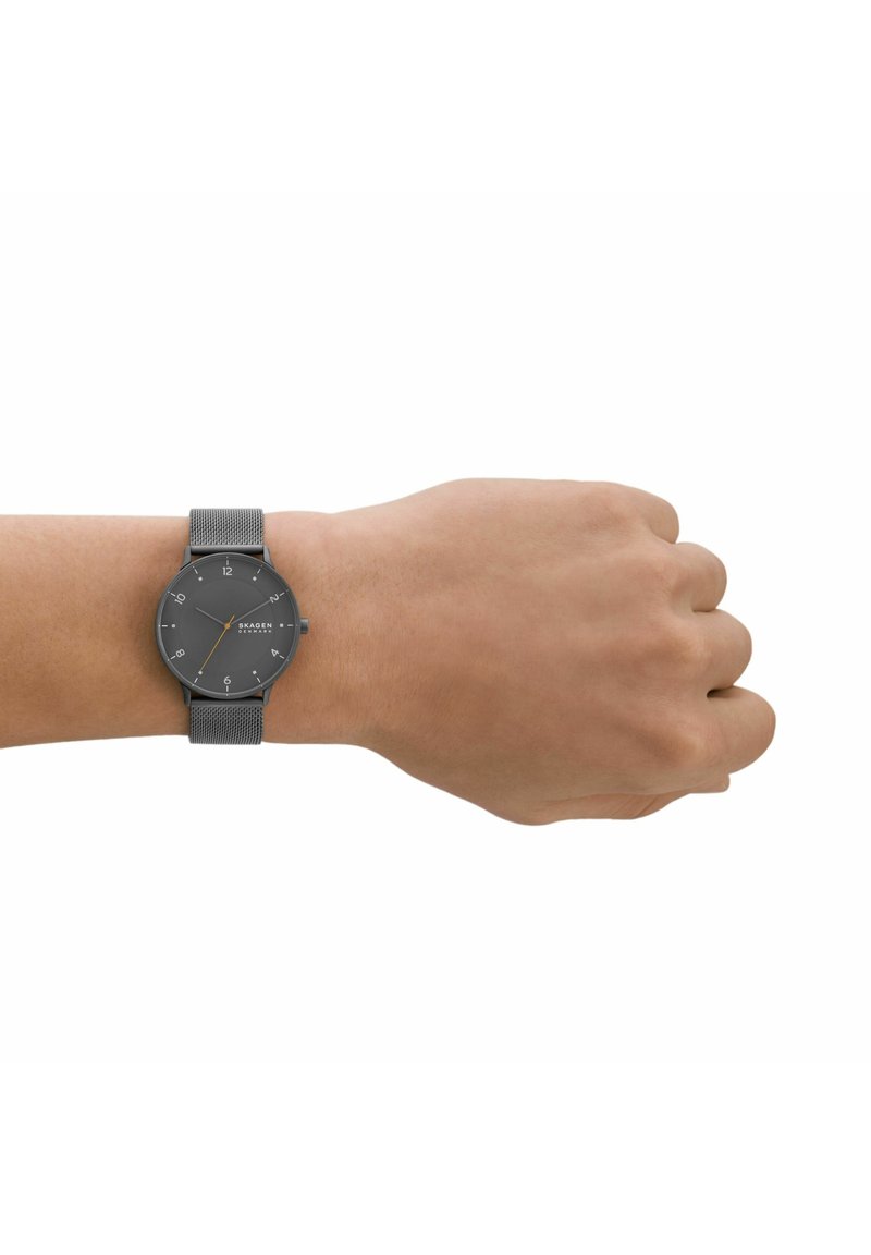 Poignet avec une montre à bracelet en maille grise avec un cadran gris foncé affichant l'heure 10:08 et le logo "Skagen Denmark" sur le cadran.