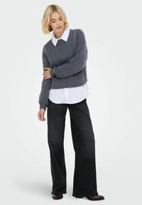 Frau mit kurzen blonden Haaren, die einen grauen Strickpullover über einem weißen Hemd, schwarze weite Jeans und dunkle Schuhe trägt, steht mit verschränkten Armen.