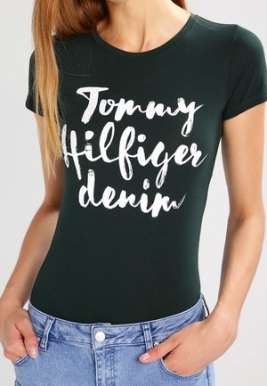 Kobieta ubrana w dopasowany ciemnozielony t-shirt z białym napisem „Tommy Hilfiger denim” i jasnoniebieskie dżinsy, z rękami w kieszeniach.