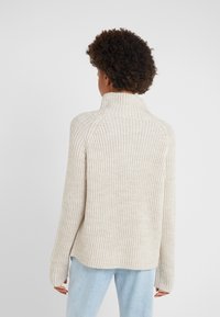 Beige knitted sweater with a tall collar, ribbed texture, and a relaxed fit. Features dropped shoulders and a slight high-low hem.