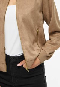 Pruun seemisnahast pommitajate jope lukuga ees, soonikkoes varrukaotstega ja lukuga küljetaskuga. Kantud valge topi ja mustade kõrge vöökohaga pükste kohal.