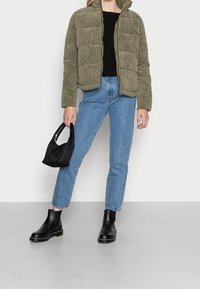 Giacca puffer in velluto a coste verde oliva con colletto alto, abbinata a jeans a vita alta blu e stivali neri alla caviglia, mentre tiene una borsa nera.