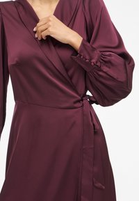 Burgundy wrap dress in smooth satin with long puff sleeves, featuring button accents and a tie waist detailing.