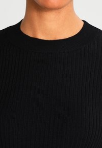 Svart, ribbad stickad topp med rund halsringning och textilstruktur som visar vertikala linjer och en slät yta.