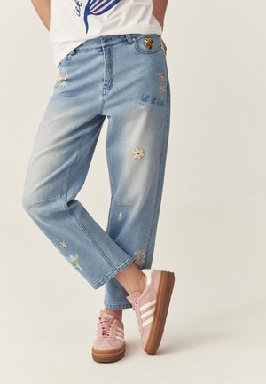 Personne portant un jean bleu clair avec une broderie florale colorée et des baskets roses à rayures blanches, debout les jambes croisées.