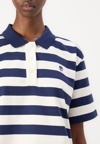 Polo con righe orizzontali blu navy e bianche, dotato di colletto, tre bottoni e un piccolo logo ad albero sul lato sinistro del petto.