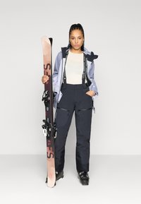 Skikleding met een lichtpaarse jas met capuchon, zwarte skibroek met zakken, en een pair ski's met bindingen, verticaal vastgehouden.