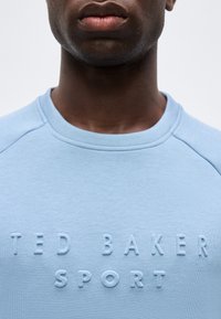 Άνδρας που φοράει ανοιχτό μπλε φούτερ με ανάγλυφο κεντημένο κείμενο «Ted Baker Sport» στο στήθος, ορατό από τον λαιμό έως το πάνω μέρος του κορμού.
