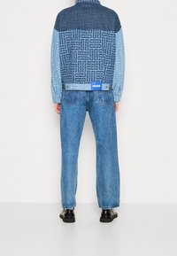 Koszula dżinsowa z niebiesko-czarnym wzorem graficznym, z logo "HUGO" i klasycznym kołnierzem, zestawiona z jasnoniebieskimi dżinsami i czarnymi butami.