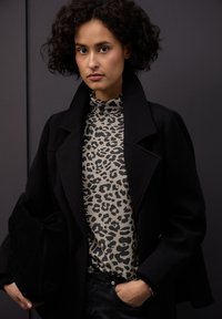 Czarny wełniany płaszcz na beżowym i czarnym leopardzim golfie. Modelka trzyma czarną zamszową kopertówkę na tle ciemnym.